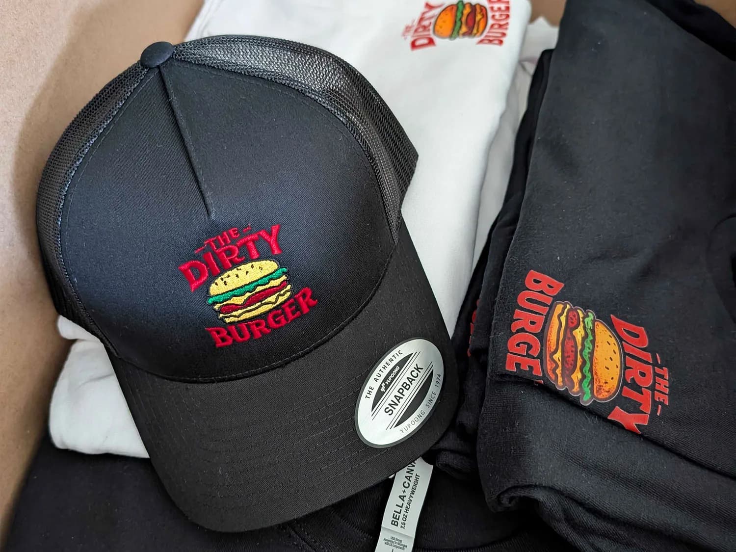 Custom Merch For Your Business Black trucker cap with an embroidered Dirty Burger logo beside black and white apparel with matching printed logos.