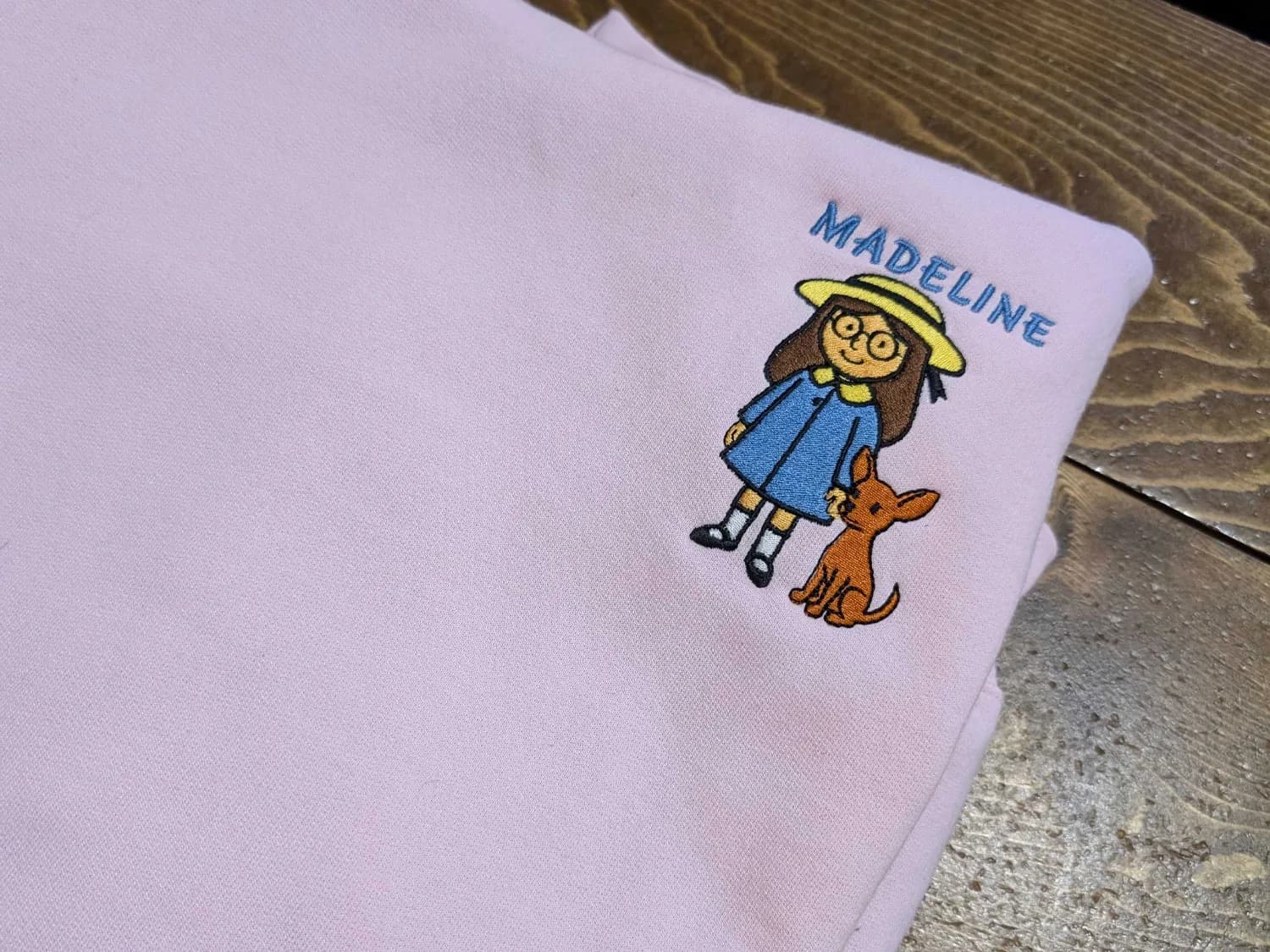 Personalized Hoodie with Character Embroidery Lavender hoodie with blue “Madeline” lettering embroidered above a girl in a yellow hat beside a brown dog.