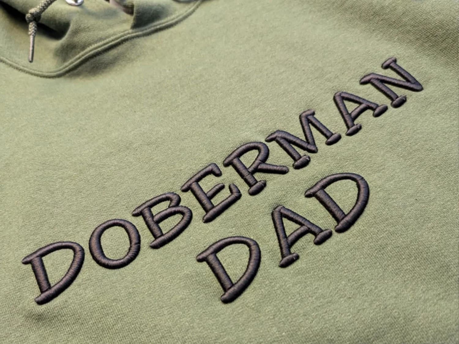 Custom Hoodie with 3D Puff Embroidery Close-up of an olive green hoodie with raised black “Doberman Dad” lettering in 3D puff embroidery.